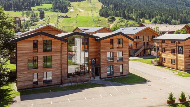 A modern wooden lodge with multiple rooms, a central glass entrance, and a parking area.