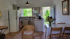 A compact kitchen with a refrigerator, microwave, and coffee maker. There is a dining table with chairs and a wicker chair with a cushion. A window provides a view of greenery outside. - Independent apartment, - 4 people (Reillanne)