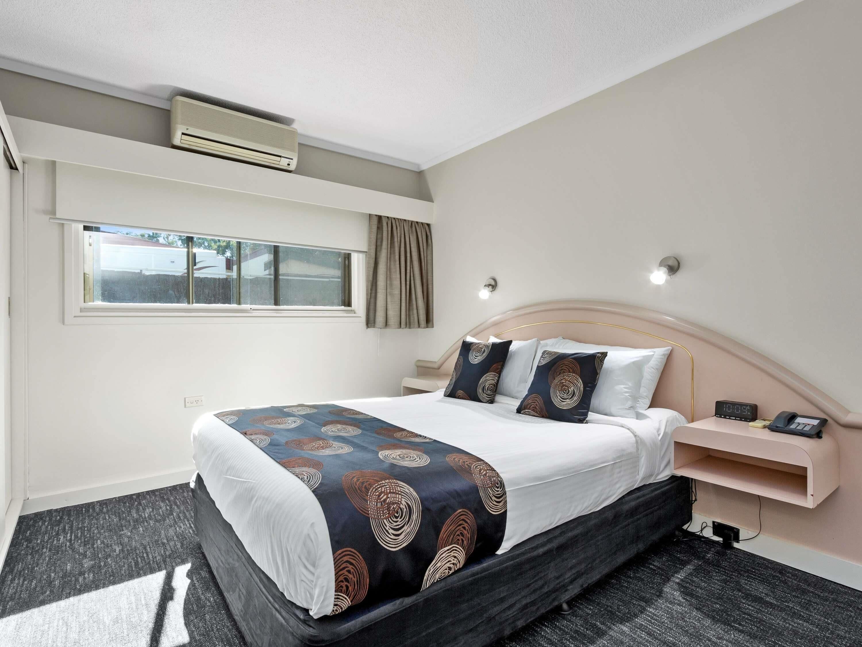 A hotel room with a bed, bedside table, nightstand, wall-mounted air conditioner, window with curtains, and a wall-mounted light fixture.