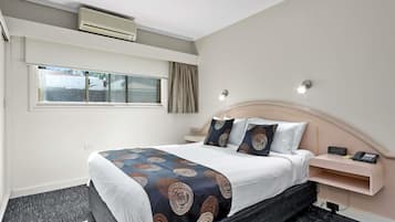 A hotel room with a bed, bedside table, nightstand, wall-mounted air conditioner, window with curtains, and a wall-mounted light fixture.
