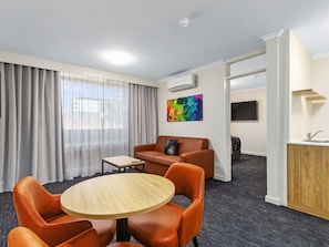 A hotel room with a round wooden table, orange leather chairs, a sofa, a TV, and an air conditioning unit.