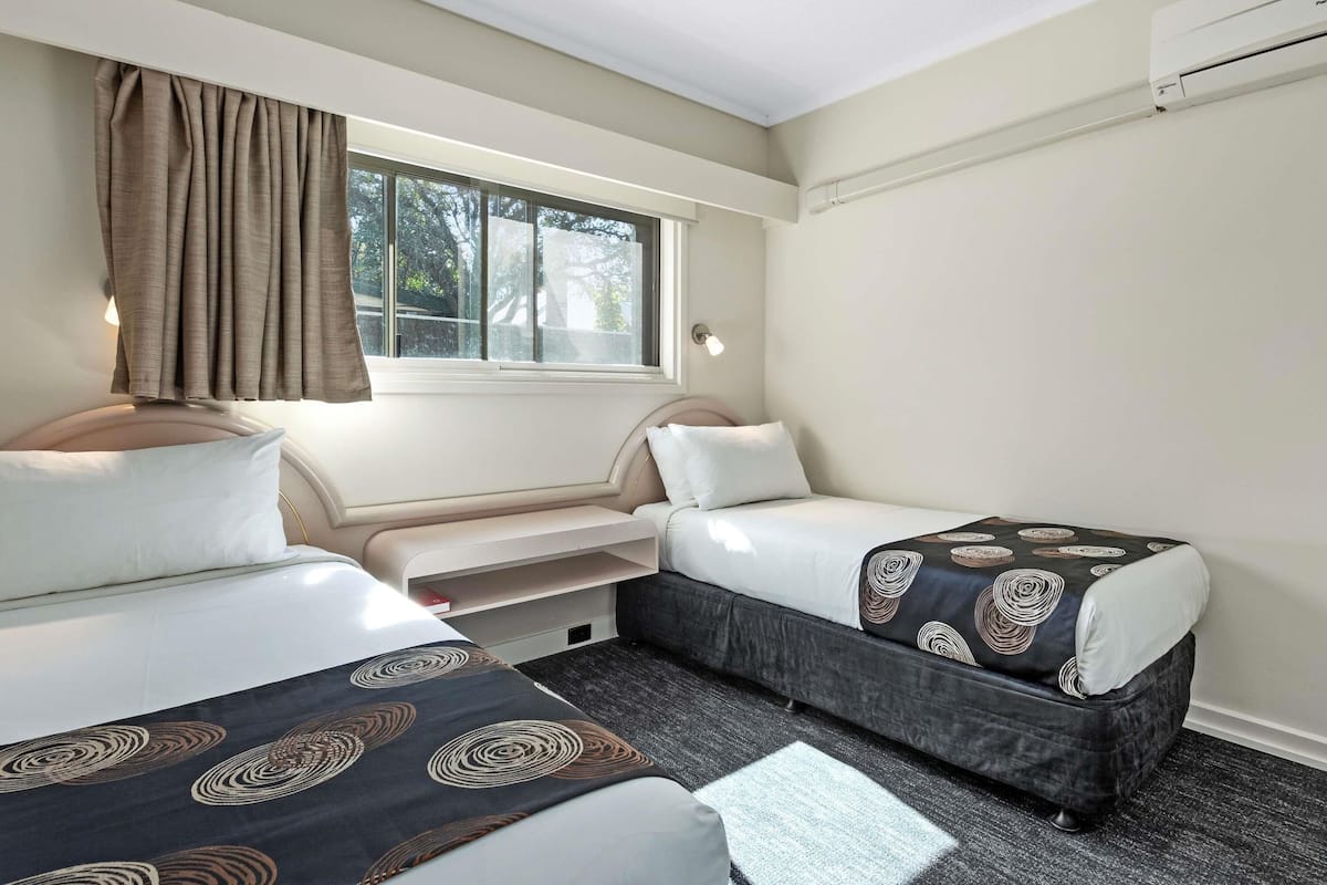 Habitación de hotel con dos camas, una ventana con cortinas, un banco empotrado y un aire acondicionado de pared.