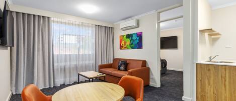 A modern hotel room with a round wooden table, orange leather chairs, a sofa, a TV, a colorful painting, and a kitchenette.
