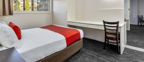 Habitación de hotel con cama, escritorio, silla, armario y ventana con vistas a edificios y árboles.