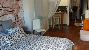 A bedroom with a patterned bedspread, a stone wall with floral decorations, a dining area with a table and chairs, and a kitchen in the background.