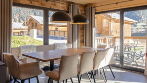 A dining area with a wooden table and chairs, large windows with curtains, and a view of wooden cabins in the background.