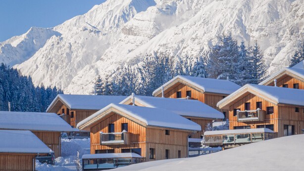 A row of wooden cabins with balconies, set against a backdrop of snow-covered mountains.