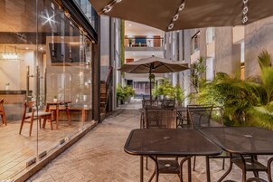 Un espacio de comedor interior y exterior con paredes de vidrio, mesas y sillas de metal, y un paraguas grande.