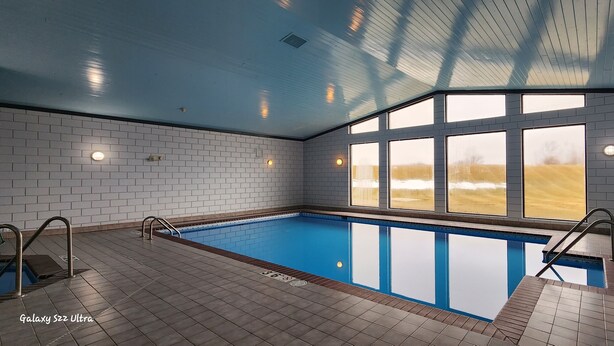 An indoor swimming pool with clear blue water, tiled walls, and large windows offering a view of a field. - The Oasis Hotel & Bar (Sheboygan Falls)