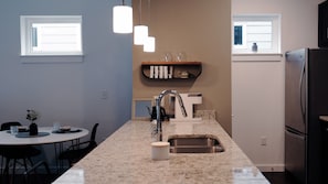 A modern kitchen with a granite countertop, stainless steel sink, and a built-in oven. There are two windows providing natural light and a shelf above the sink holding cups and a teapot.