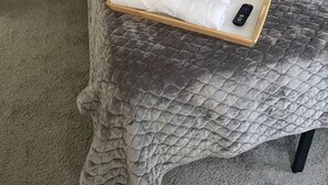 Un lit bien fait, recouvert d’une courtepointe matelassée grise, avec deux oreillers et un plateau en bois sur lequel sont posés des serviettes pliées et une télécommande.