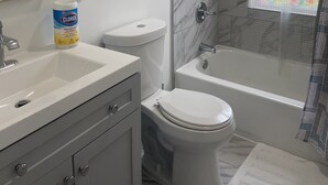 Une salle de bain avec des toilettes, un lavabo et une baignoire. On y trouve un miroir, une serviette et une plante sur l’étagère.