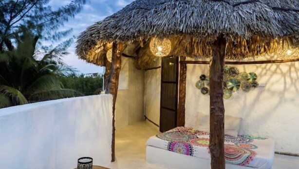 Uma cabana com telhado de palha, uma cama e um lustre pendurado.
