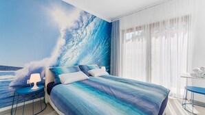 Une chambre avec un grand lit, une table de chevet, une lampe, une chaise et une vue sur l’océan à travers de fines rideaux.