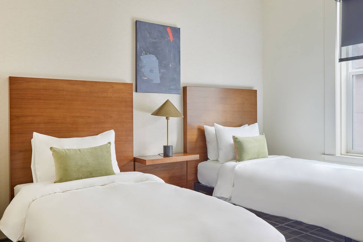 A hotel room with two beds, a nightstand with a lamp, a painting on the wall, and a window.