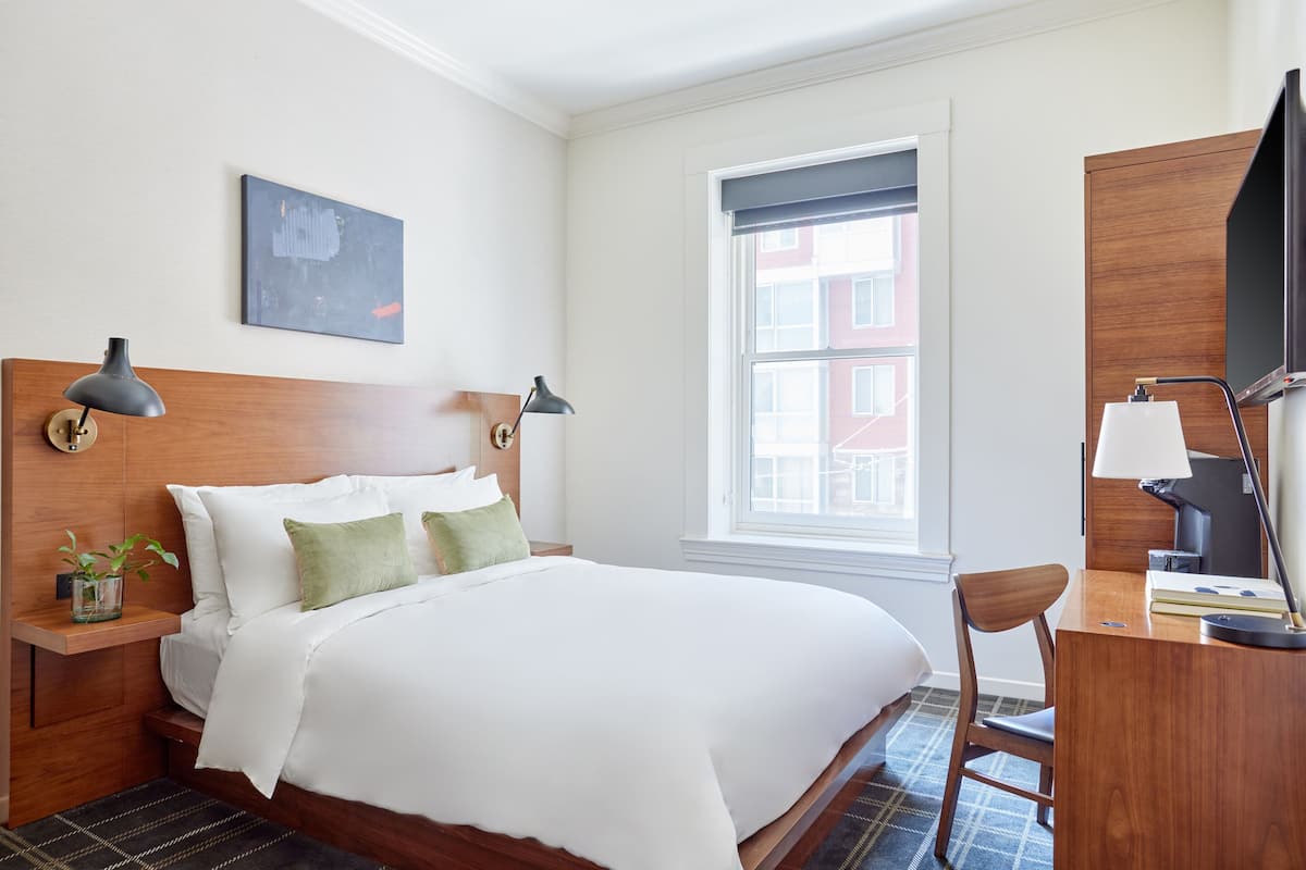 A hotel room with a large bed, two bedside lamps, a desk with a chair, a television, and a window with a view of buildings.