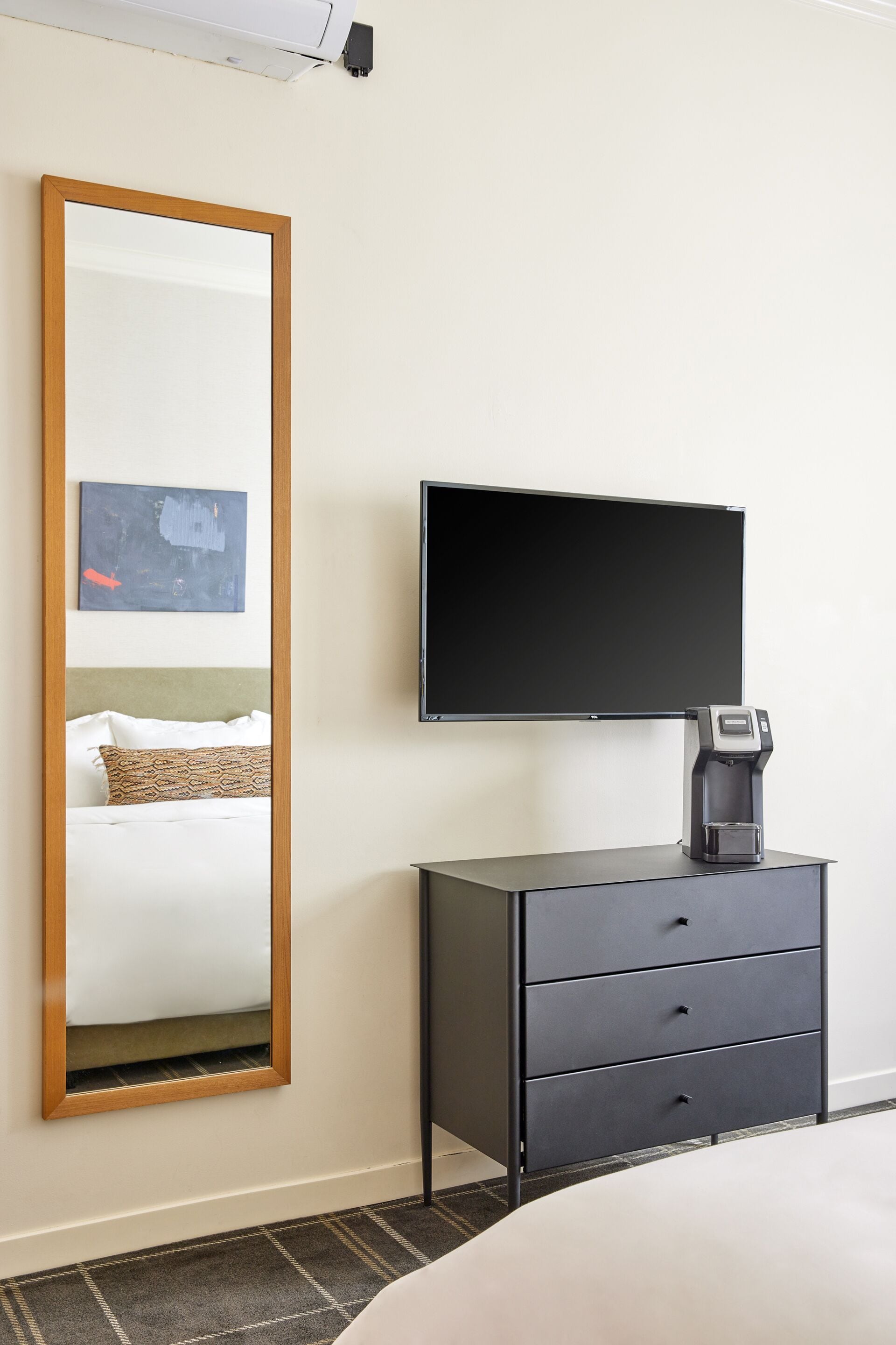 A hotel room with a large mirror, a flat-screen TV mounted on the wall, a coffee maker on a black cabinet, and a bed with white bedding.