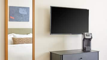A hotel room with a large mirror, a flat-screen TV mounted on the wall, a coffee maker on a black cabinet, and a bed with white bedding.