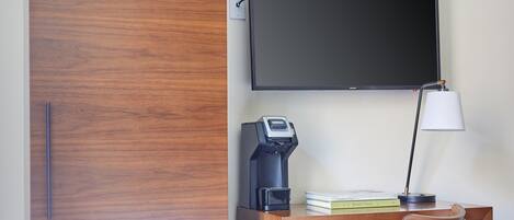A hotel room with a wooden wardrobe, a desk with a lamp, a coffee maker, and a flat-screen TV mounted on the wall.