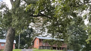 Une maison en bois rouge, dotée d’un porche, entourée d’une végétation luxuriante et d’arbres.