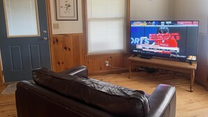 Un salon avec un canapé en cuir, un téléviseur diffusant des programmes sportifs, une porte décorée d’une étoile et un tableau encadré accroché au mur.