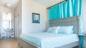 A bedroom with a bed, a nightstand, a window with curtains, a wall-mounted picture, and a bathroom with a sink and mirror. - LaCasita Ocean Front House (Aguada)