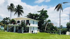 A two-story house with a covered patio, surrounded by a well-maintained lawn and palm trees. - LaCasita Ocean Front House (Aguada)