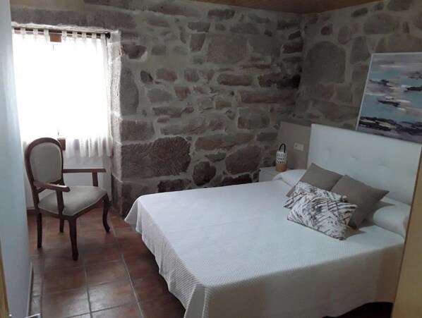 A bedroom with a stone wall, a bed with a white bedspread, a chair, and a small table. - CORUXA HOUSE (San Salvador de Poio)