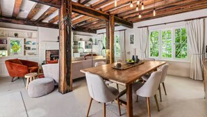 Un vaste espace de vie comprenant une table à manger en bois, des chaises blanches et un fauteuil rouge. On y trouve également une cheminée, un coin cuisine et de grandes fenêtres avec des rideaux.