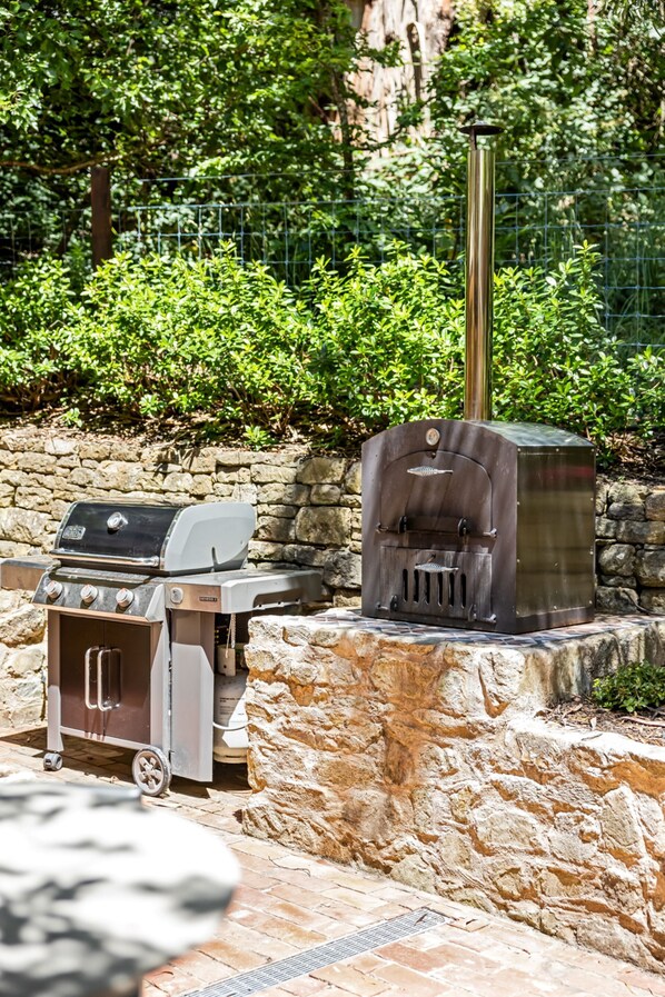 A barbecue grill and a wood-fired oven set up against a stone wall with a metal fence and greenery in the background.
