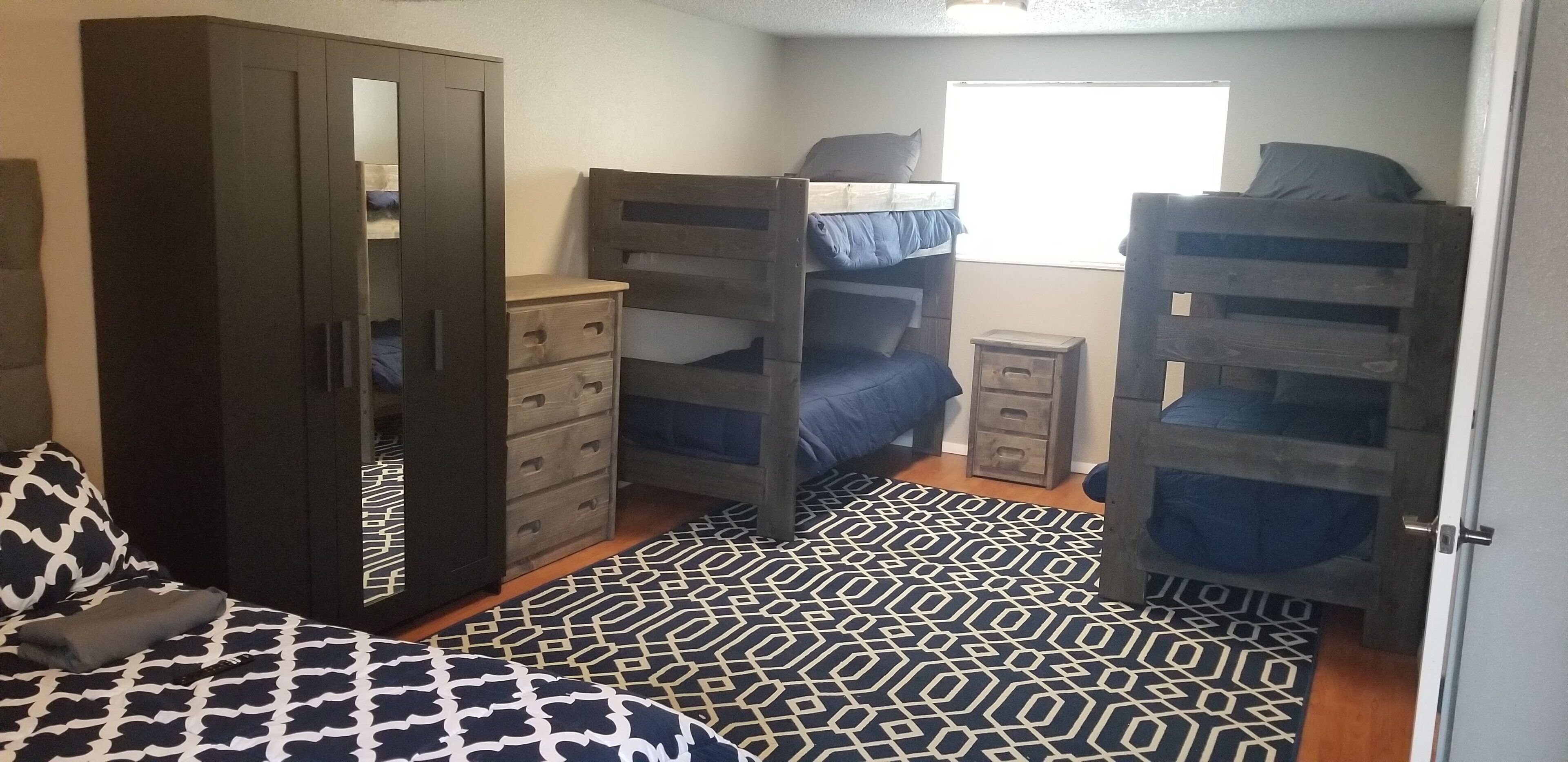 A dormitory room with bunk beds, a wardrobe, a dresser, and a patterned rug.