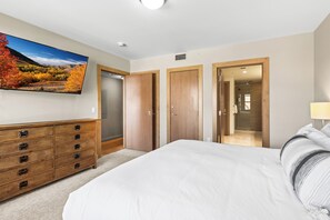 A bedroom with a large bed, a wooden dresser, a TV mounted on the wall, and a view of a forest through the window.