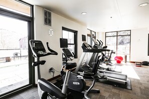 Academia com diversos equipamentos de ginástica, janelas amplas e vista para o exterior.