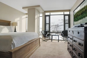 Ein Hotelzimmer mit einem großen Bett, einem Schreibtisch, zwei Stühlen, einer Kommode und einem Fenster mit Blick auf eine verschneite Landschaft.