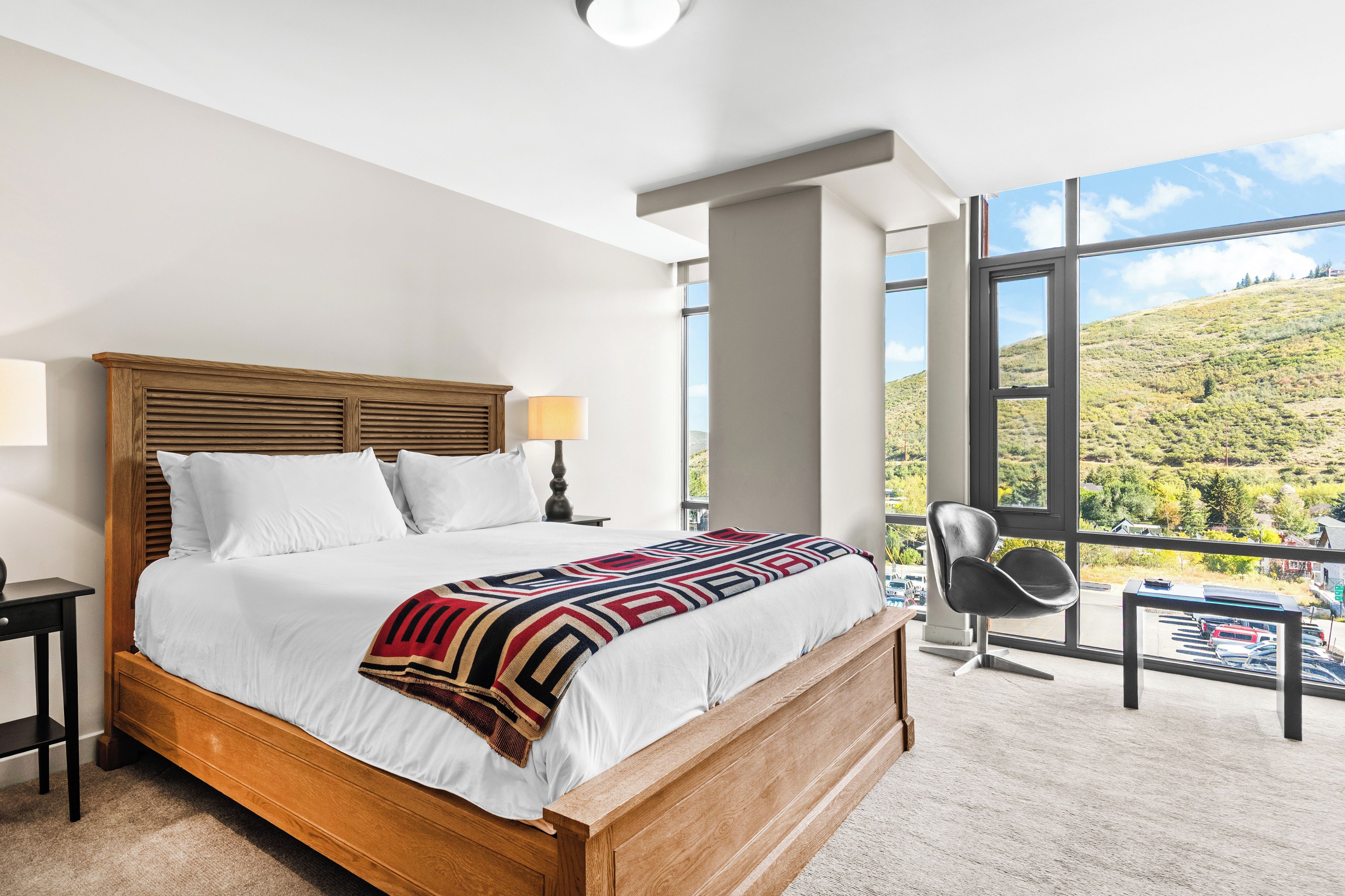 Een hotelkamer met een groot bed, uitzicht op een glooiend landschap, een moderne stoel en een nachtkastje met een lamp.