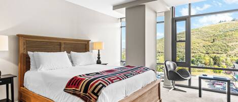 Een hotelkamer met een groot bed, uitzicht op een glooiend landschap, een moderne stoel en een nachtkastje met een lamp.