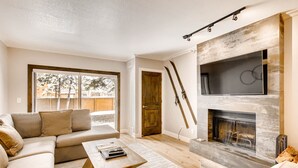 Ein modernes Wohnzimmer mit einer Eckcouch, einem Holztisch, einem Kamin und einem großen Fenster mit Blick ins Freie.