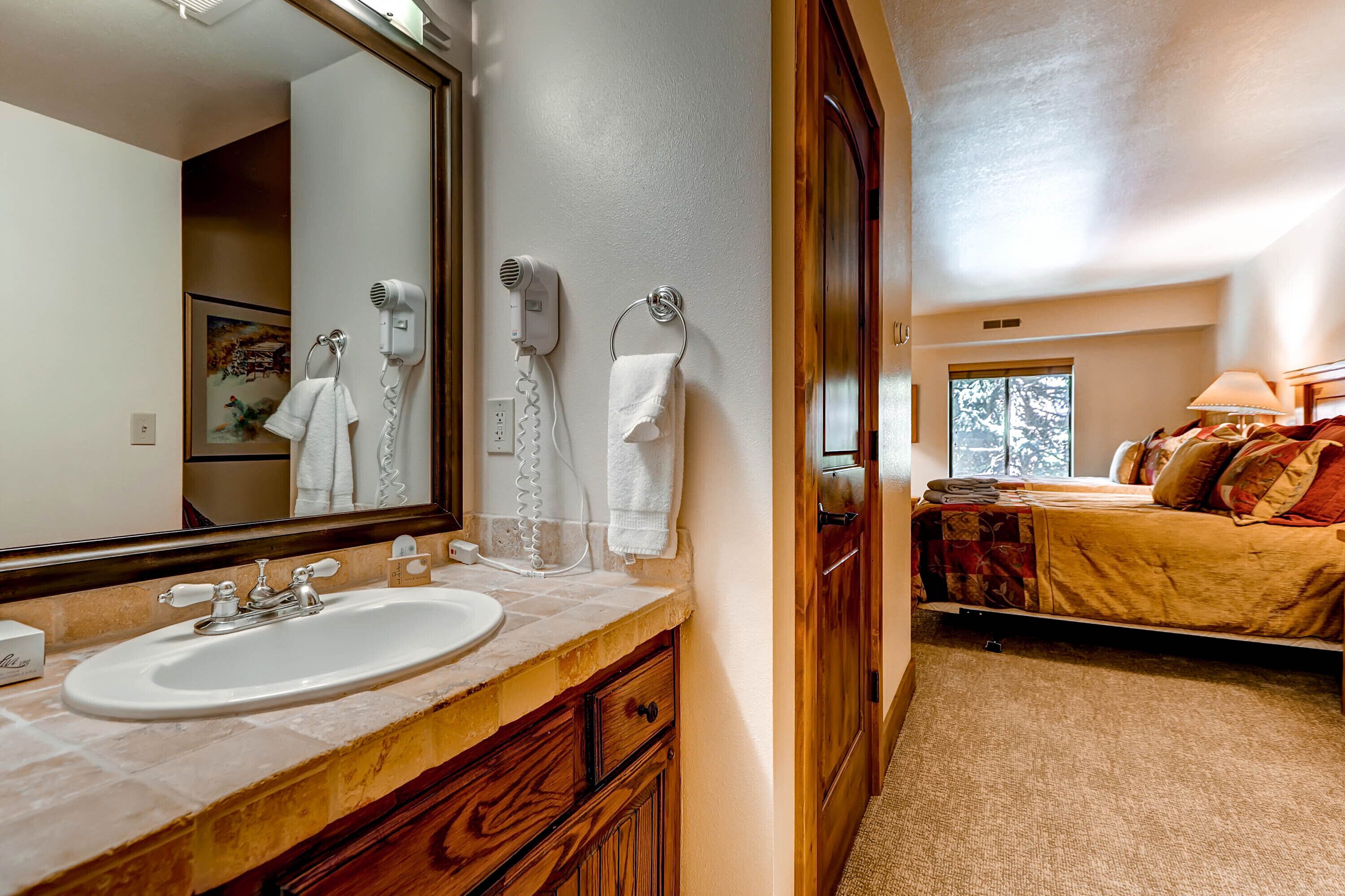 Une salle de bain équipée d’un lavabo, d’un miroir et d’un sèche-cheveux. La pièce adjacente contient un lit et une fenêtre.