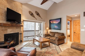 A cozy living room with a fireplace, leather furniture, a flat-screen TV, and a large window overlooking a snowy landscape.