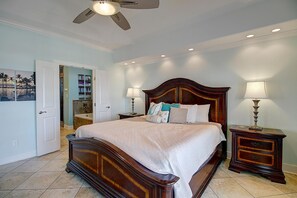 A bedroom with a large bed, two bedside tables, a ceiling fan, recessed lighting, and a door leading to another room.