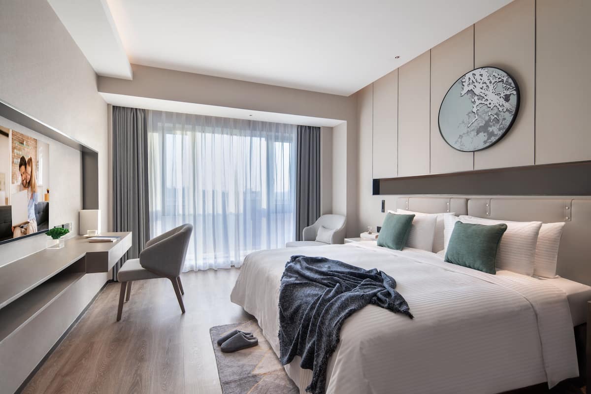 Quarto de hotel moderno com uma cama grande, uma escrivaninha, uma cadeira e uma janela ampla com cortinas leves.