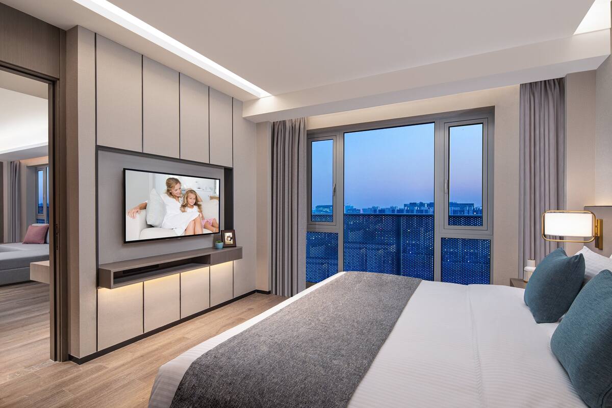 Quarto de hotel moderno com uma cama grande, televisão de tela plana na parede, vista da cidade através de amplas janelas e um espaço de estar com sofá e poltrona.