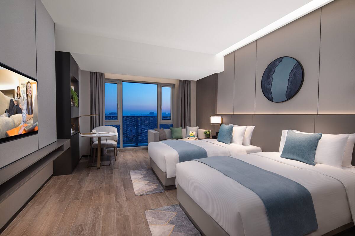 Quarto de hotel moderno com duas camas, televisão, escrivaninha e uma janela grande com vista para a cidade.