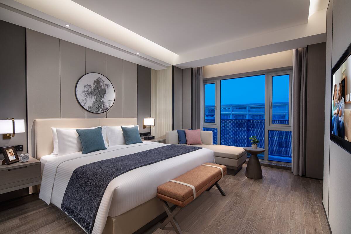 Quarto de hotel moderno com uma cama grande, área de estar e varanda com vista para a cidade.