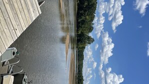 Une jetée en bois s’étendant sur une étendue d’eau calme, entourée d’une forêt dense et surplombée d’un ciel bleu clair parsemé de quelques nuages.