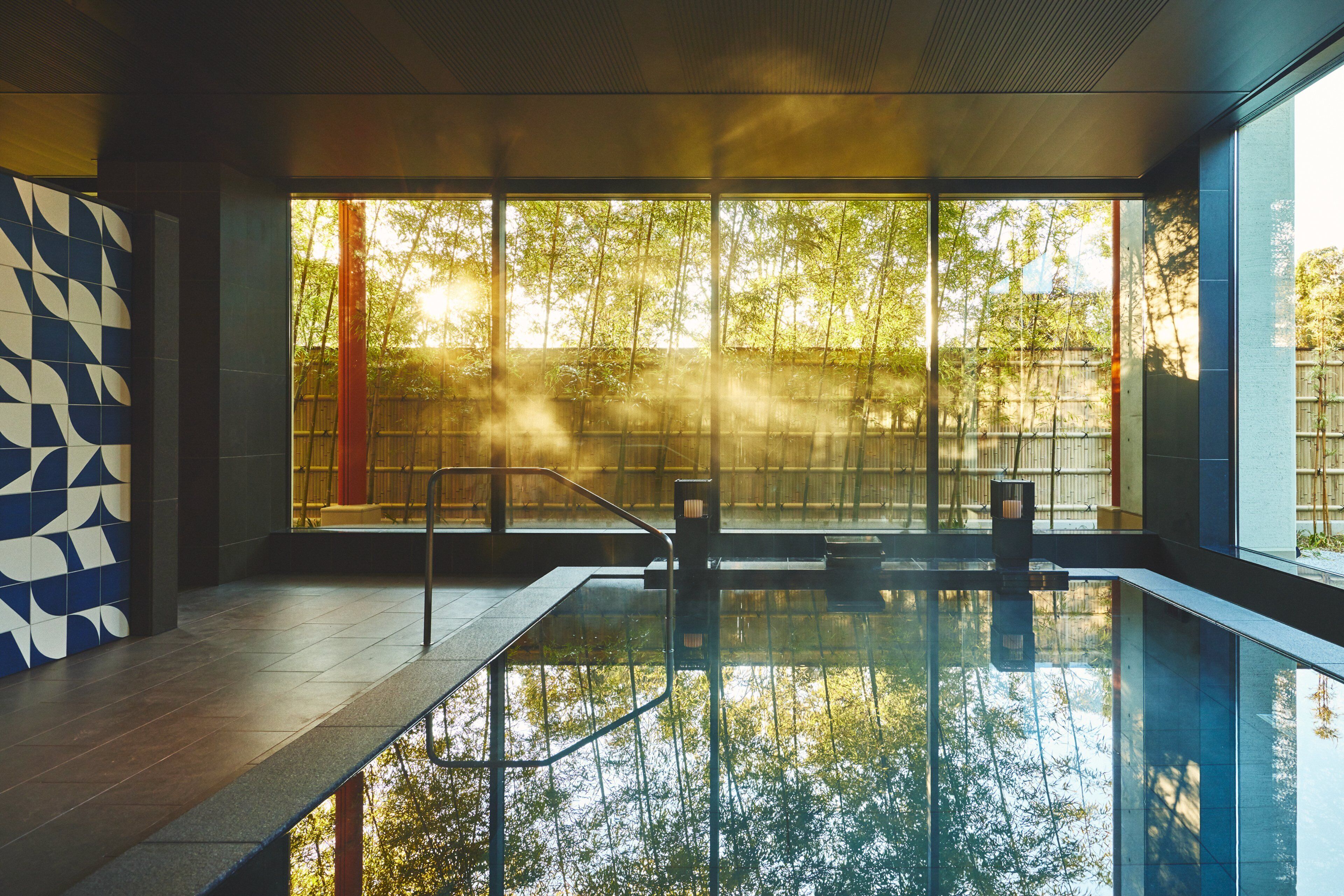 A modern indoor swimming pool with clear glass walls, a geometric tile wall, and a view of trees outside.