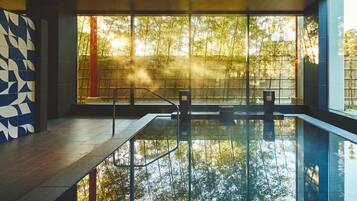 A modern indoor swimming pool with clear glass walls, a geometric tile wall, and a view of trees outside.
