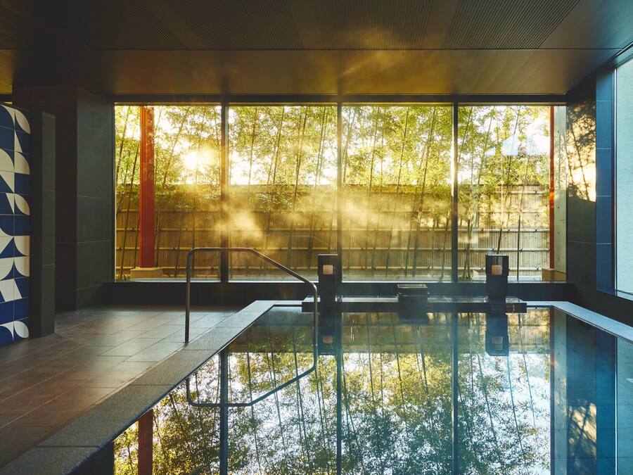 A modern indoor swimming pool with clear glass walls, a geometric tile wall, and a view of trees outside.