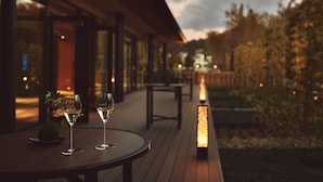 Una terraza de madera con dos copas de vino blanco, una planta en maceta y un farol encendido.