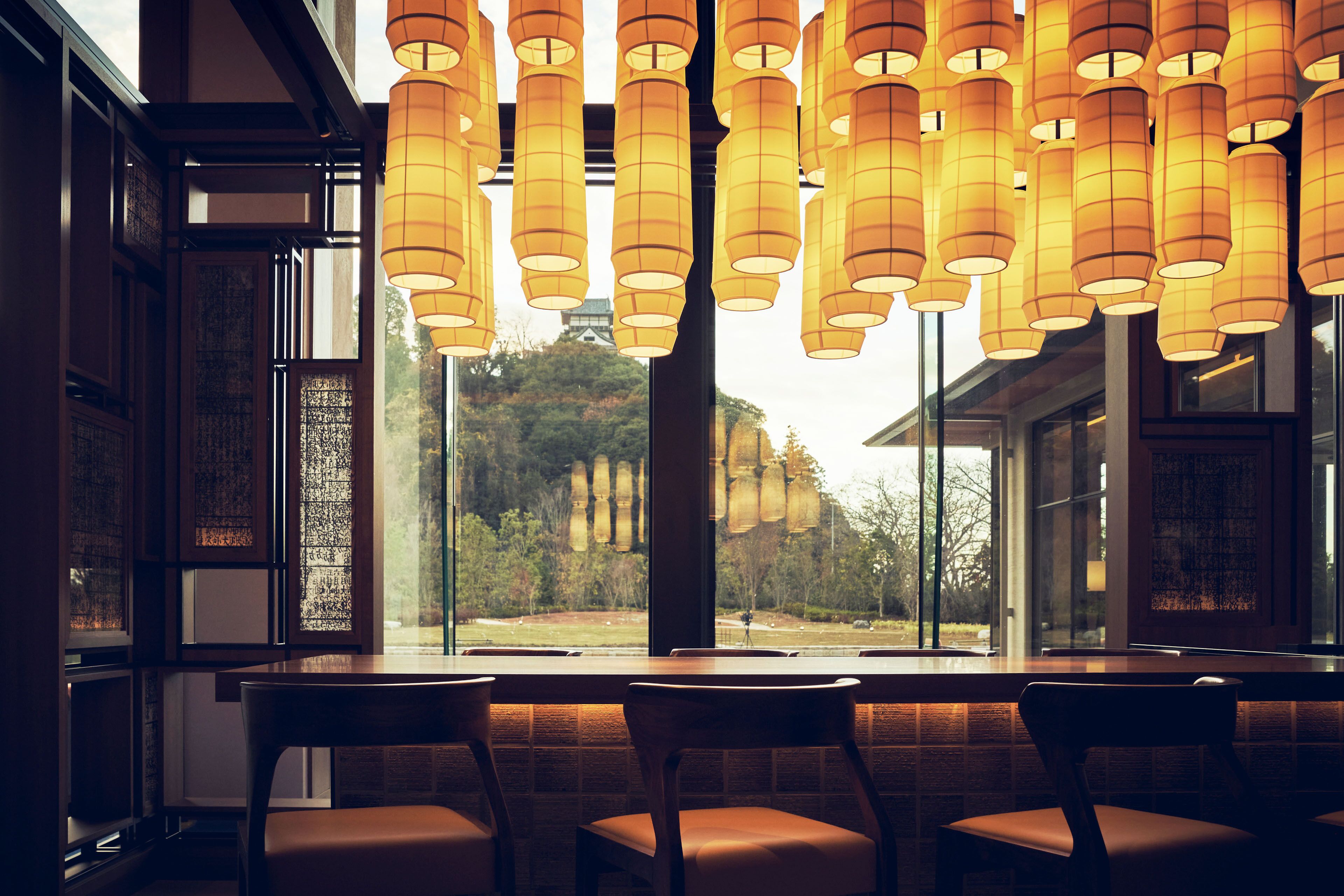 Interior de restaurante con varias mesas y sillas, amplias ventanas con vistas a una colina y árboles, y lámparas colgantes de estilo farol.